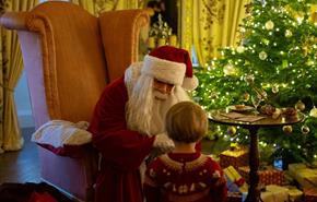Father Christmas with a child infront of a decorated Christmas tree surrounded by presents.